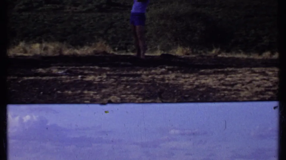 1969 Kenya 8mm frame showing rhinoceroses on dusty savanna with shrubs, sparse trees, blue sky, and cropped human legs above.