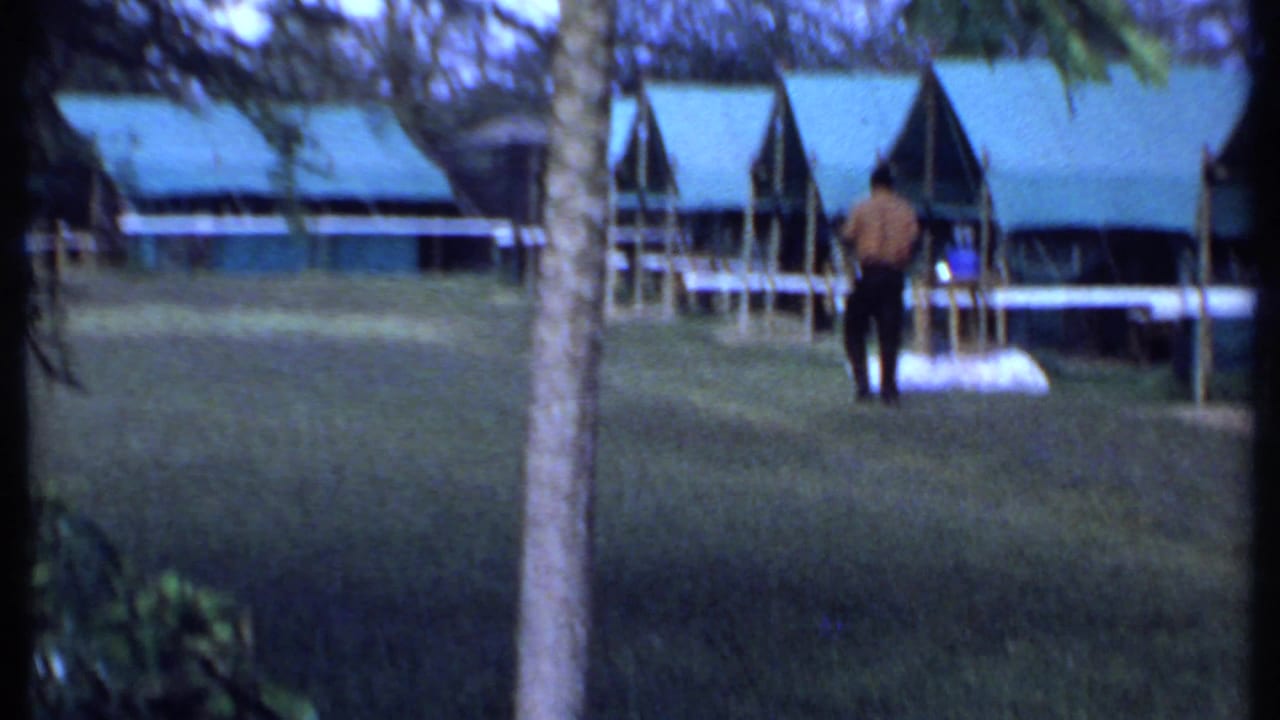 A distant adult stands on a grassy camp area in Kenya, with low green-roofed safari buildings and trees in the background.