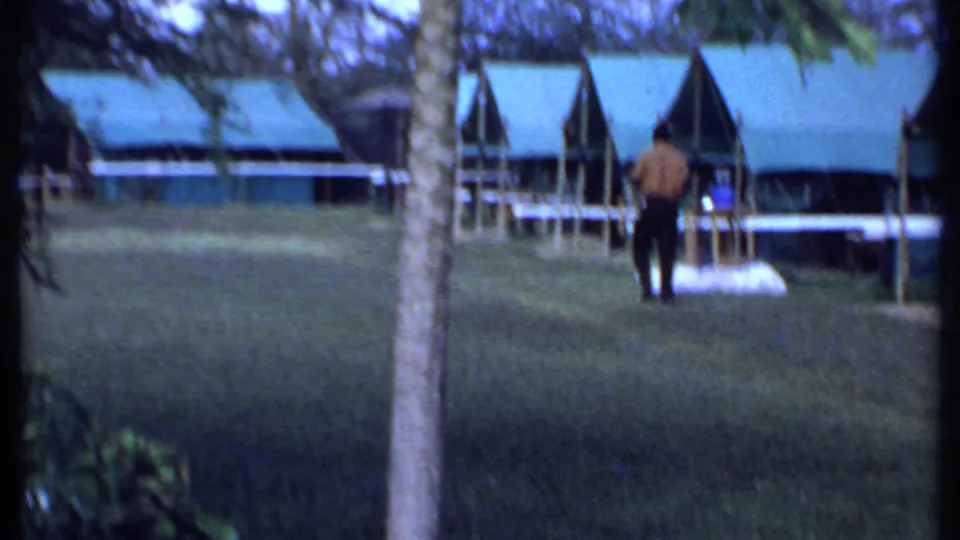 A distant adult stands on a grassy camp area in Kenya, with low green-roofed safari buildings and trees in the background.