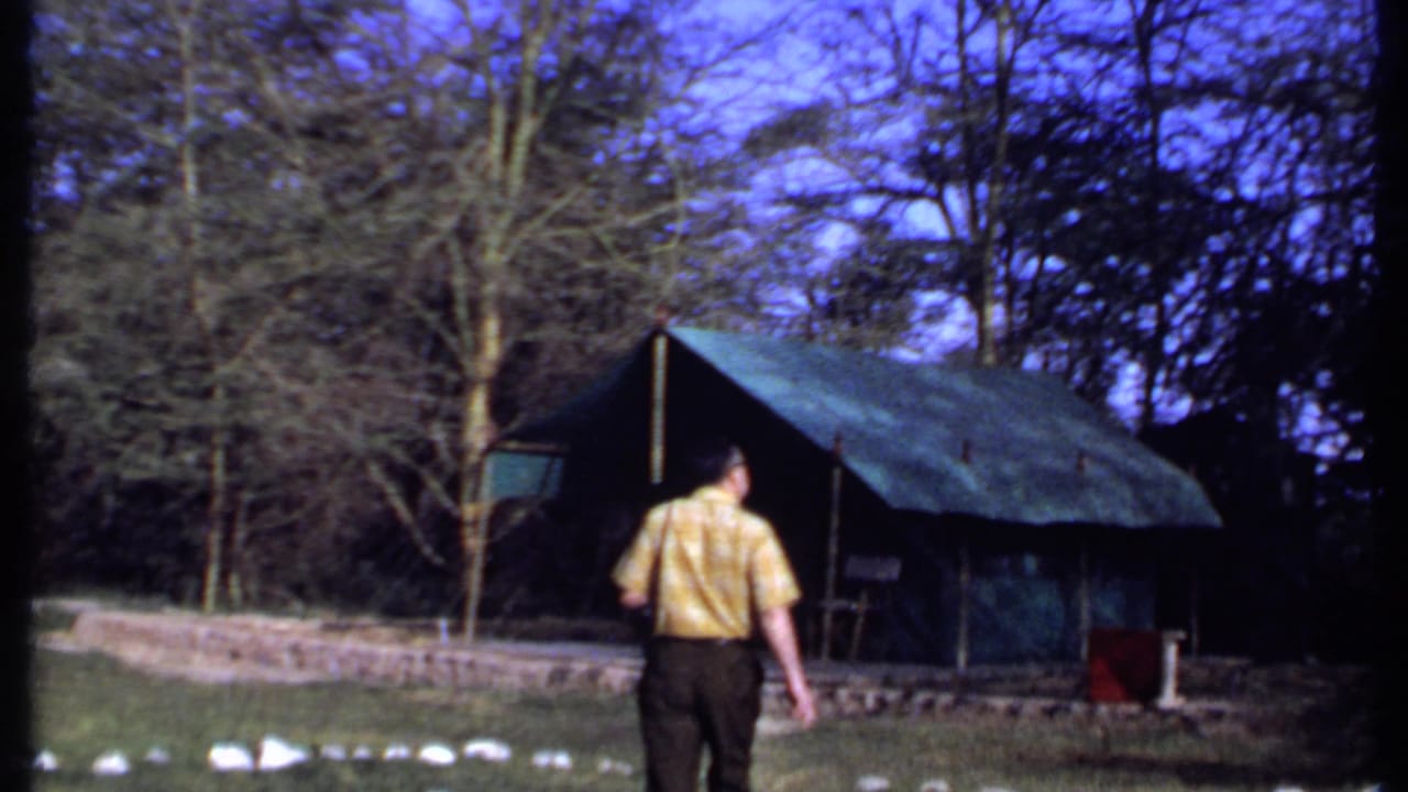 Man in a yellow shirt walks across grass toward a green canvas tent among trees.