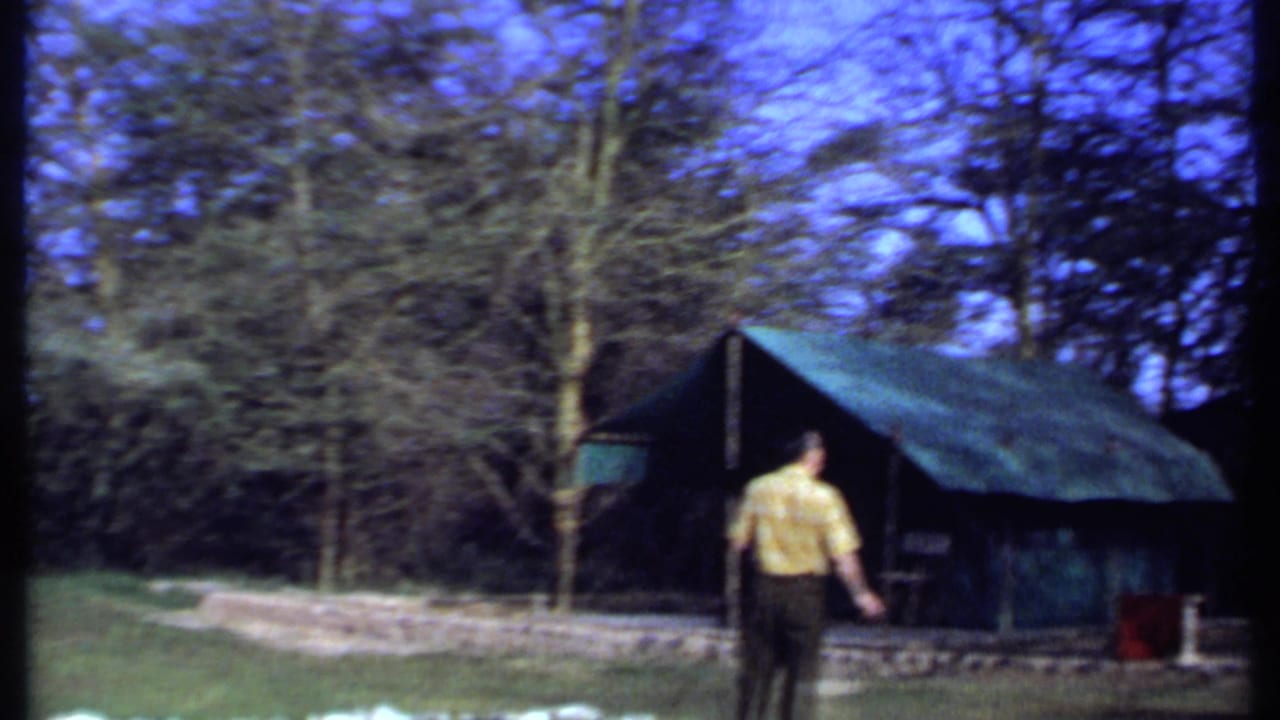 A closer view shows the man from behind near the green tent, with bare trees and a low stone border around the camp area.