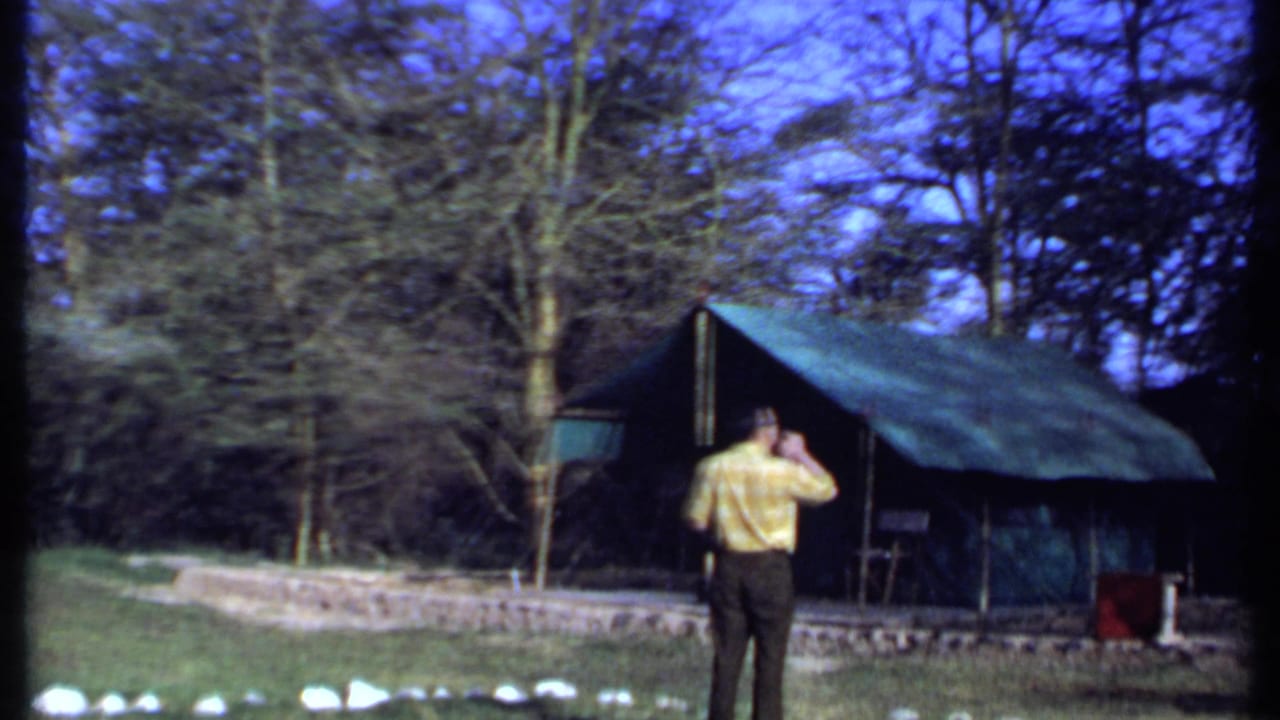 At the end of the Kenya 1969 clip, the man raises a hand near his head while facing the green canvas tent in the wooded camp.