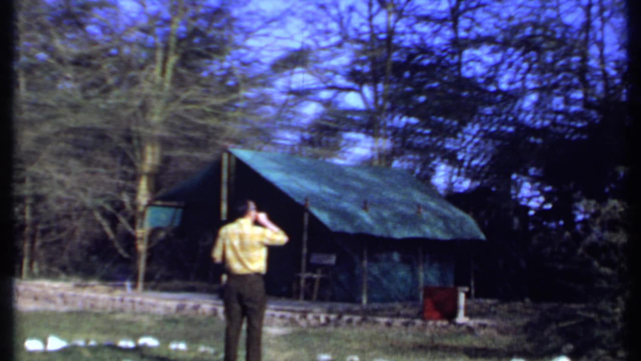Senior man in a yellow shirt stands in a wooded yard near green-roofed shelters.