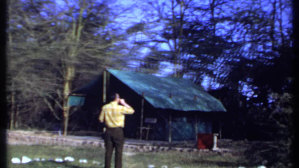 1969 Kenya 8mm frame showing a senior man in a yellow shirt shading his eyes near rustic green-roofed shelters and trees.