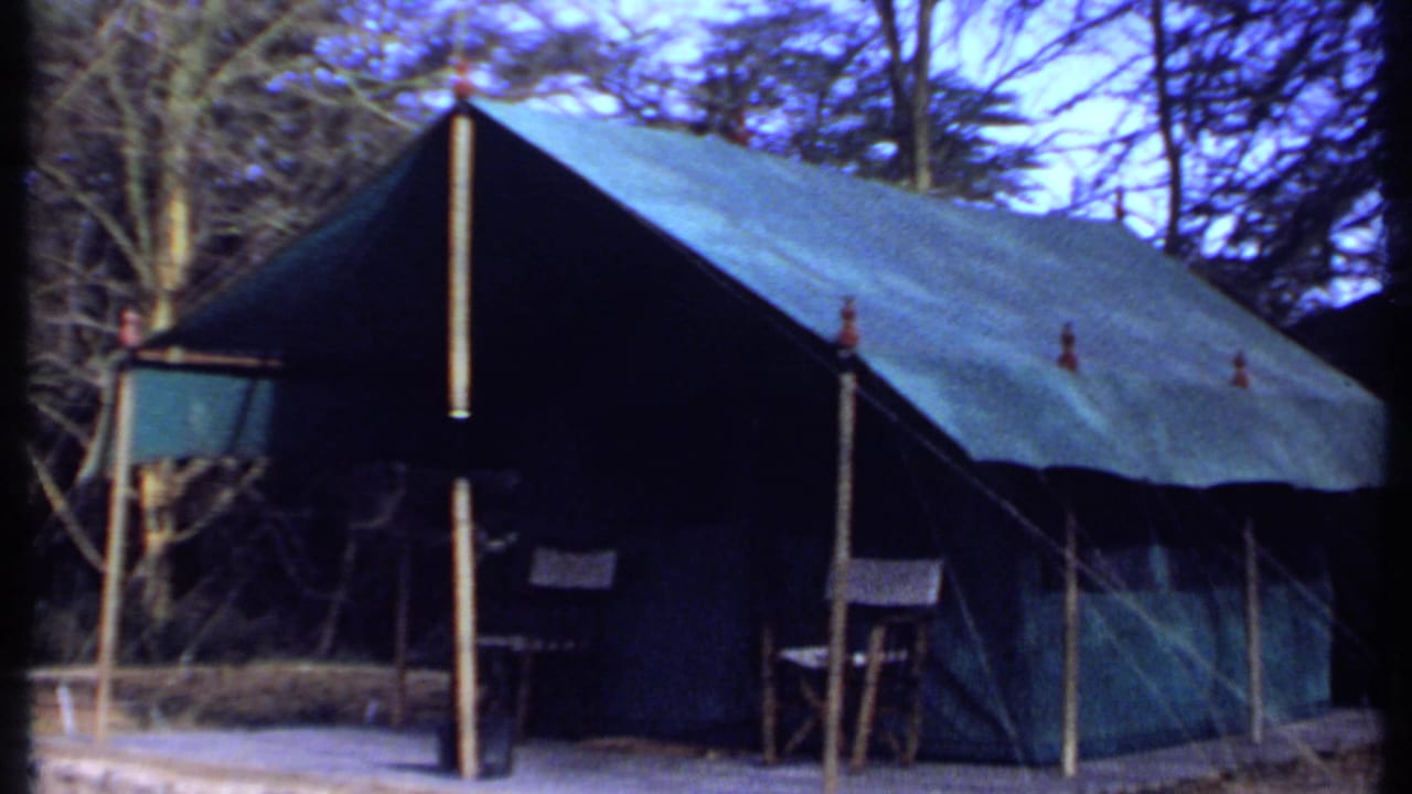 1969 Kenya frame showing a large green canvas tent set on a platform, with poles, open sides, folding chairs and surrounding trees.