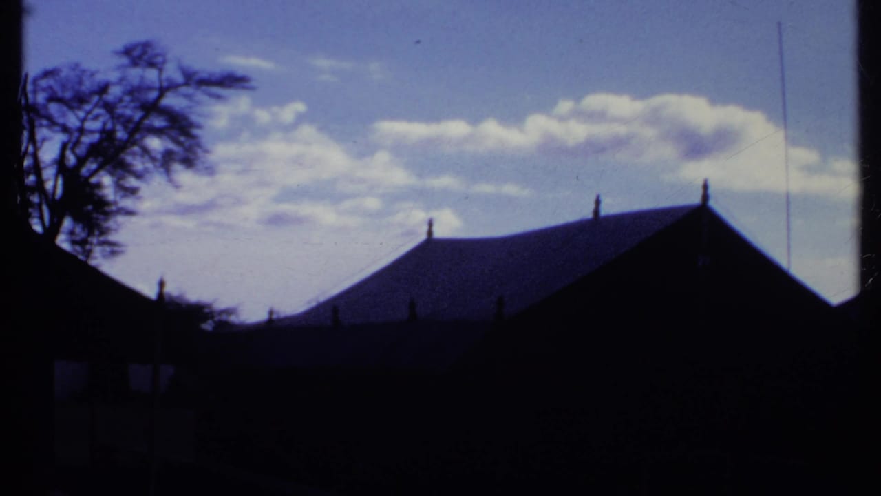 1969 Kenya home-movie frame showing dark thatched-roof structures silhouetted against a vivid blue sky.