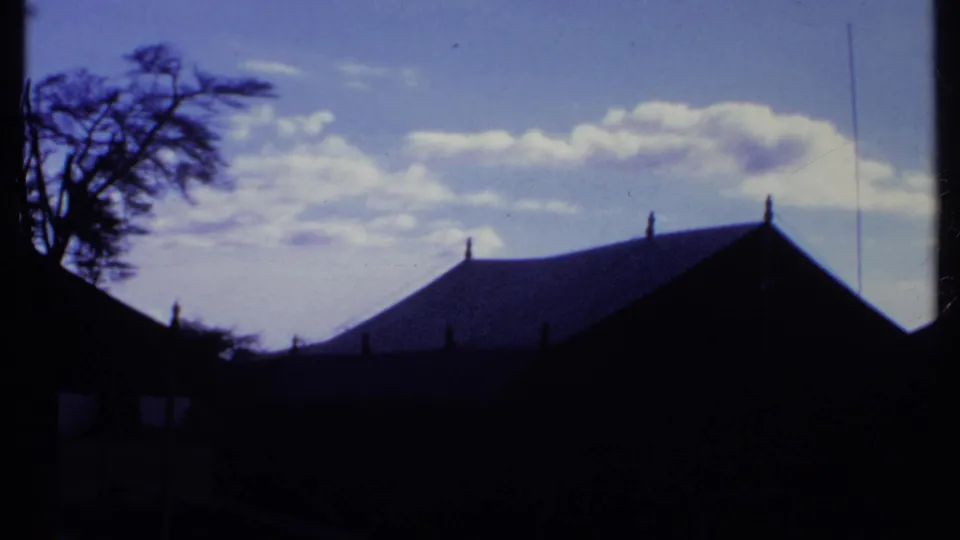 1969 Kenya home-movie frame showing dark thatched-roof structures silhouetted against a vivid blue sky.