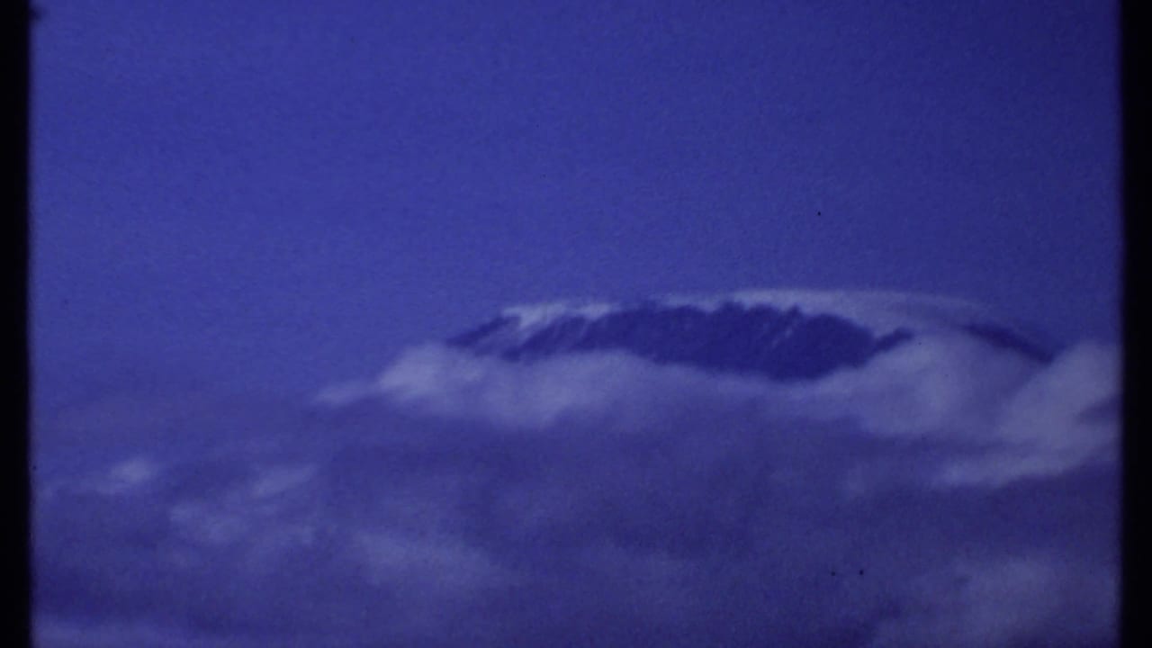 1969 Kenya 8mm frame showing a distant snow-capped mountain peak partly obscured by cloud beneath a deep blue sky.