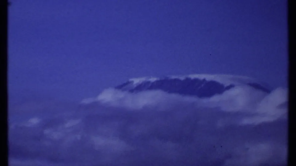 1969 Kenya 8mm frame showing a distant snow-capped mountain peak partly obscured by cloud beneath a deep blue sky.
