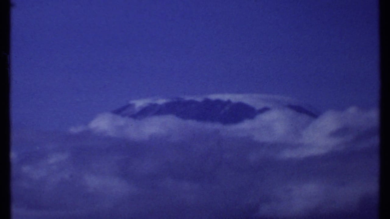 1969 Kenya 8mm frame of a cloud-wrapped snowy mountain summit seen from a distance in saturated blue light.
