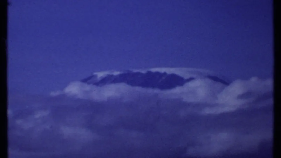1969 Kenya 8mm frame of a cloud-wrapped snowy mountain summit seen from a distance in saturated blue light.