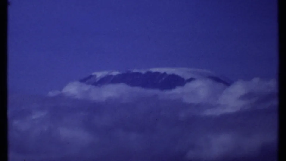 1969 Kenya 8mm frame showing a distant snow-capped mountain summit partly wrapped in clouds against a saturated blue sky.