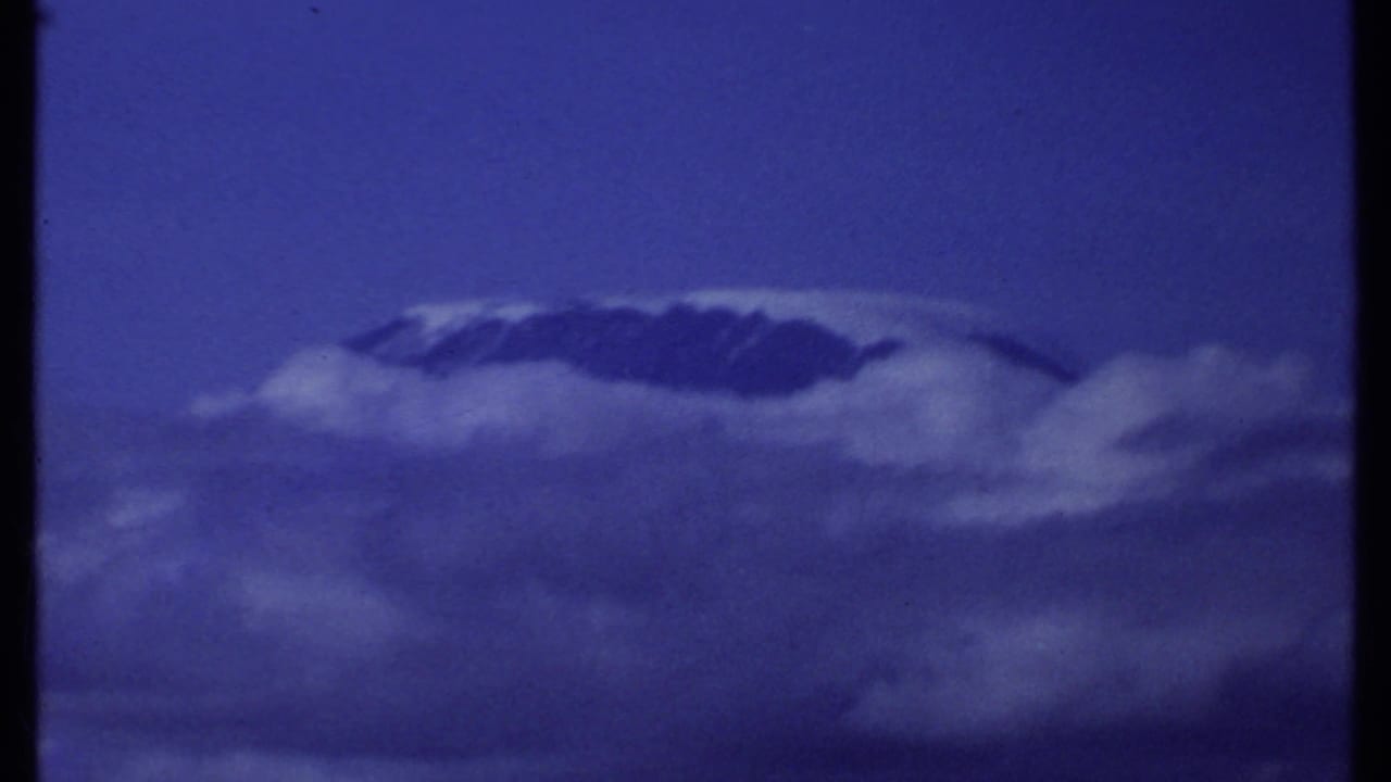 1969 Kenya 8mm aerial view of a snow-topped mountain ridge rising through cloud layers in a blue sky.