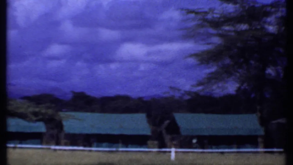 1969 Kenya home-movie view of green-roofed buildings, trees, white fence, distant hills, and dramatic blue sky.