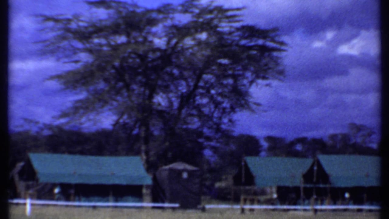 1969 Kenya 8mm frame with trees, low green-roofed structures, white fencing, and open rural ground.