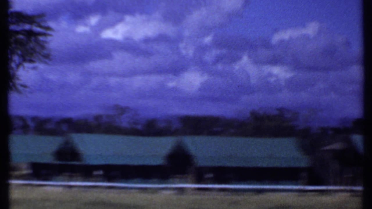 1969 Kenya landscape frame showing a long green-roofed building, white fence, distant trees, and heavy blue sky.