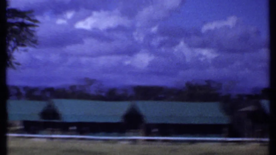 1969 Kenya landscape frame showing a long green-roofed building, white fence, distant trees, and heavy blue sky.