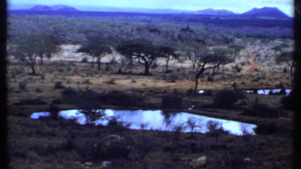 1969 Kenya 8mm frame showing open savanna, scattered trees, small reflective pools, and distant mountain forms under blue haze.