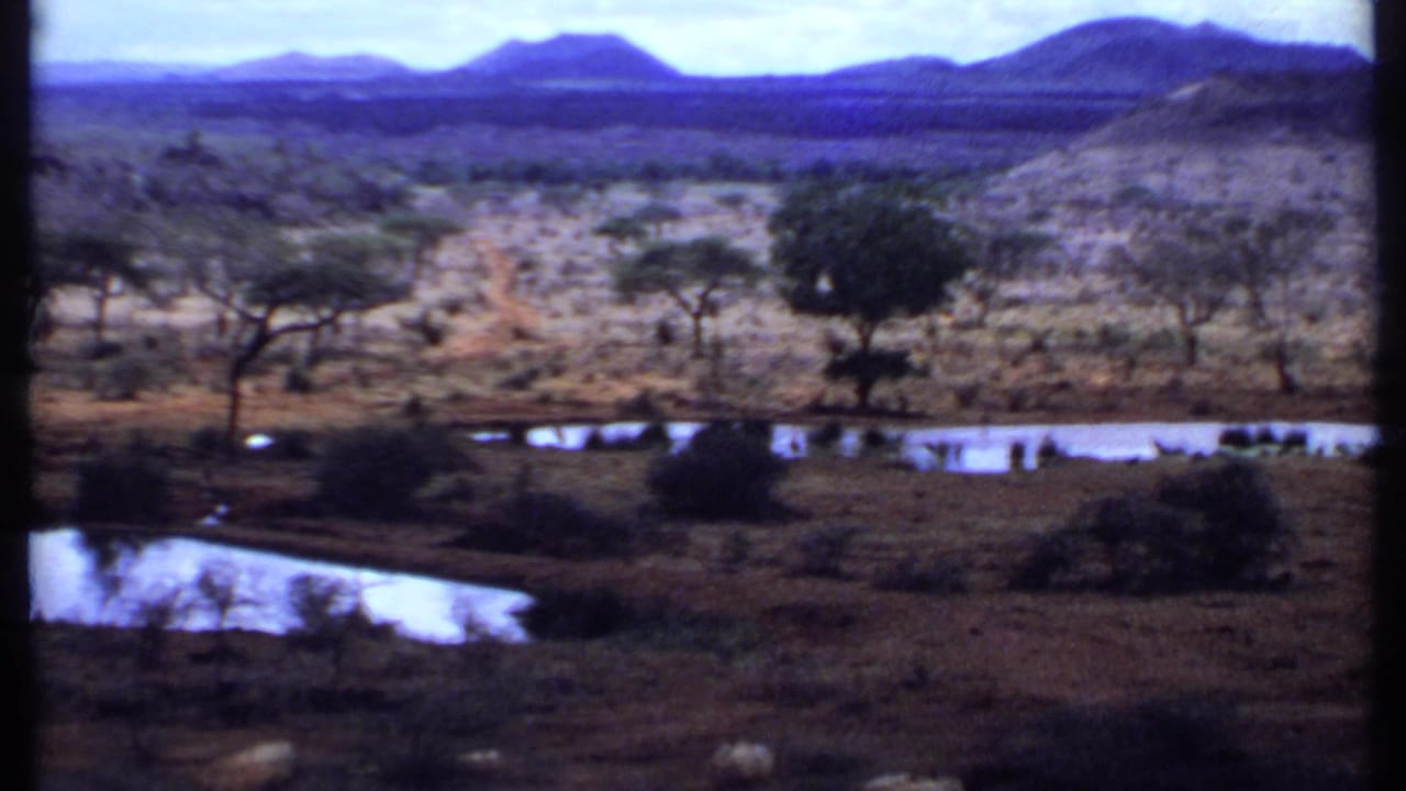 1969 Kenya 8mm frame showing dry open terrain, scattered trees, bright water reflections, and layered mountains in the distance.