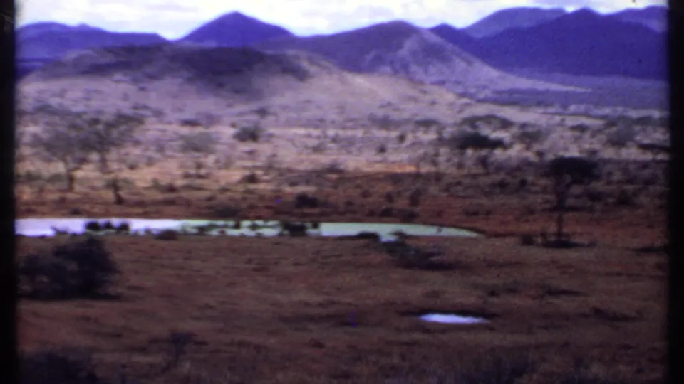 1969 Kenya home-movie frame looking across a dry plain toward a small lake, sparse trees, and blue-toned mountain ridges.