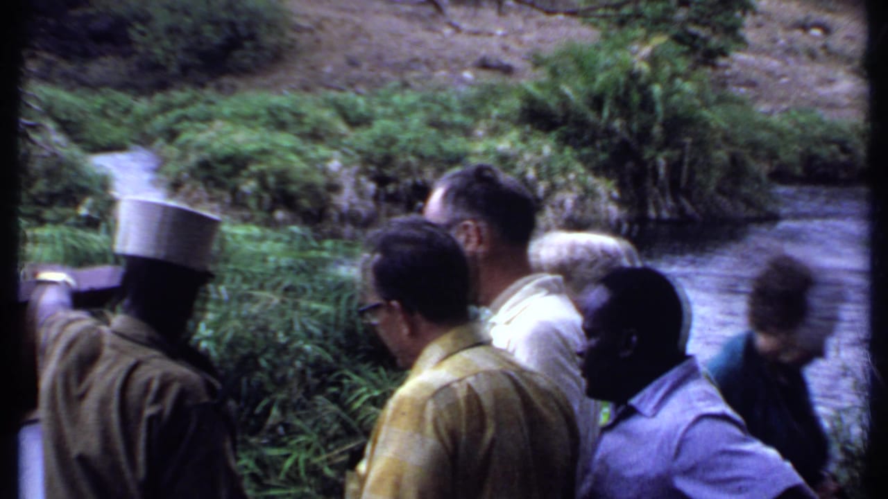 Men on a riverbank watch the safari scene, including a guide-like figure in a tall hat, with water and dense plants behind them.
