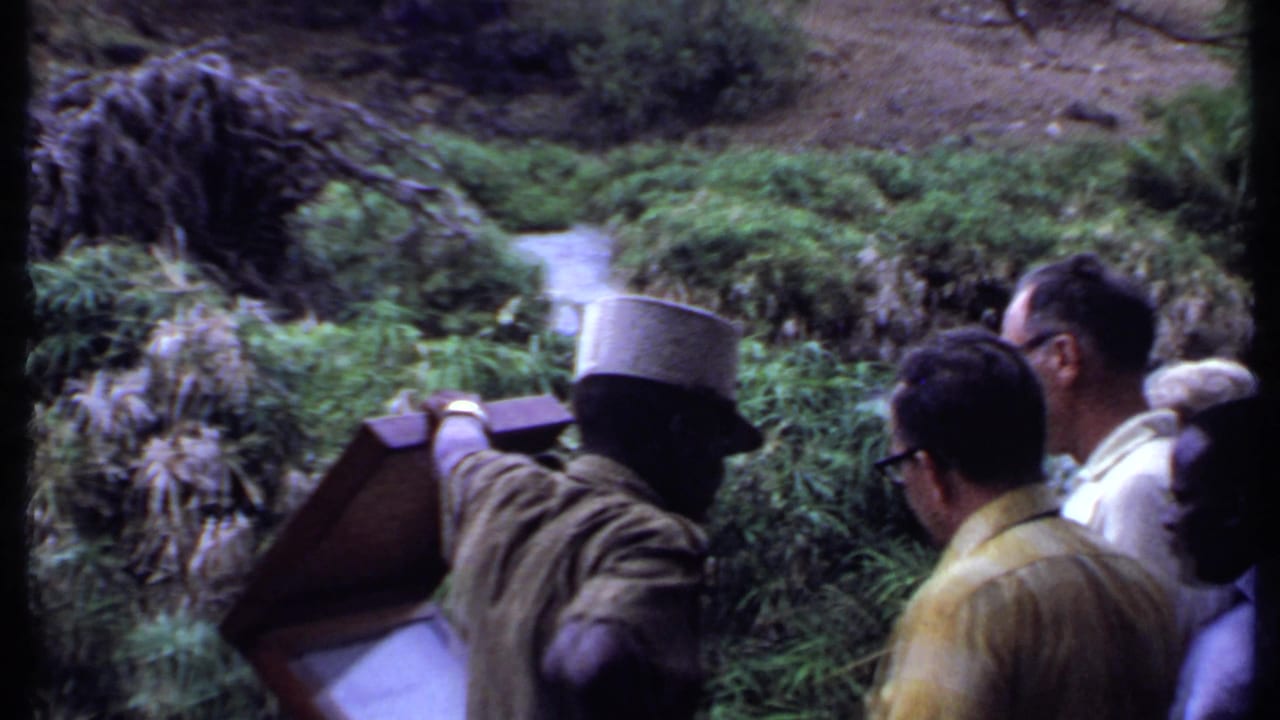 A guide-like figure gestures across a river as several men watch, framed in grainy 1969 Kenya safari home-movie footage.