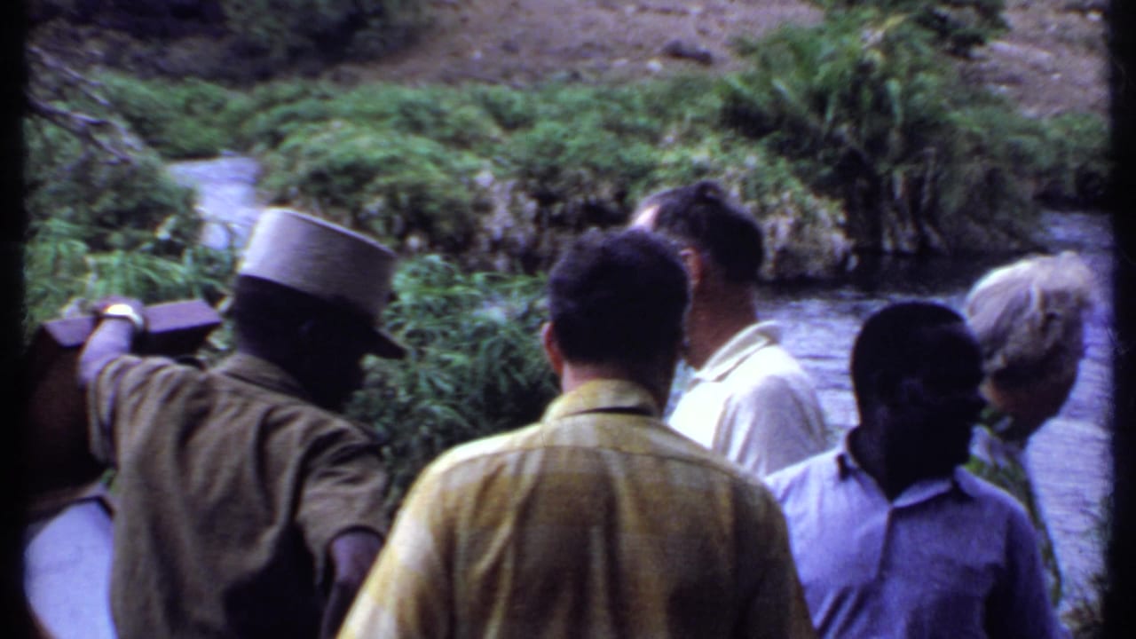 Adult tourists cluster around a guide and signboard near a small river, with lush vegetation filling the background in 1969 Kenya footage.