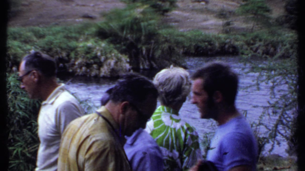 Tourists beside a river in Kenya appear to examine the ground or water area, with dense bankside plants visible in the 1969 home-movie frame.