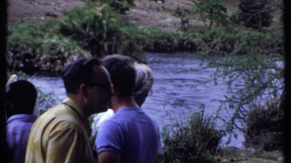 1969 Kenya home-movie frame showing tourists gathered by a flowing river, with trees and rocky banks in the background.