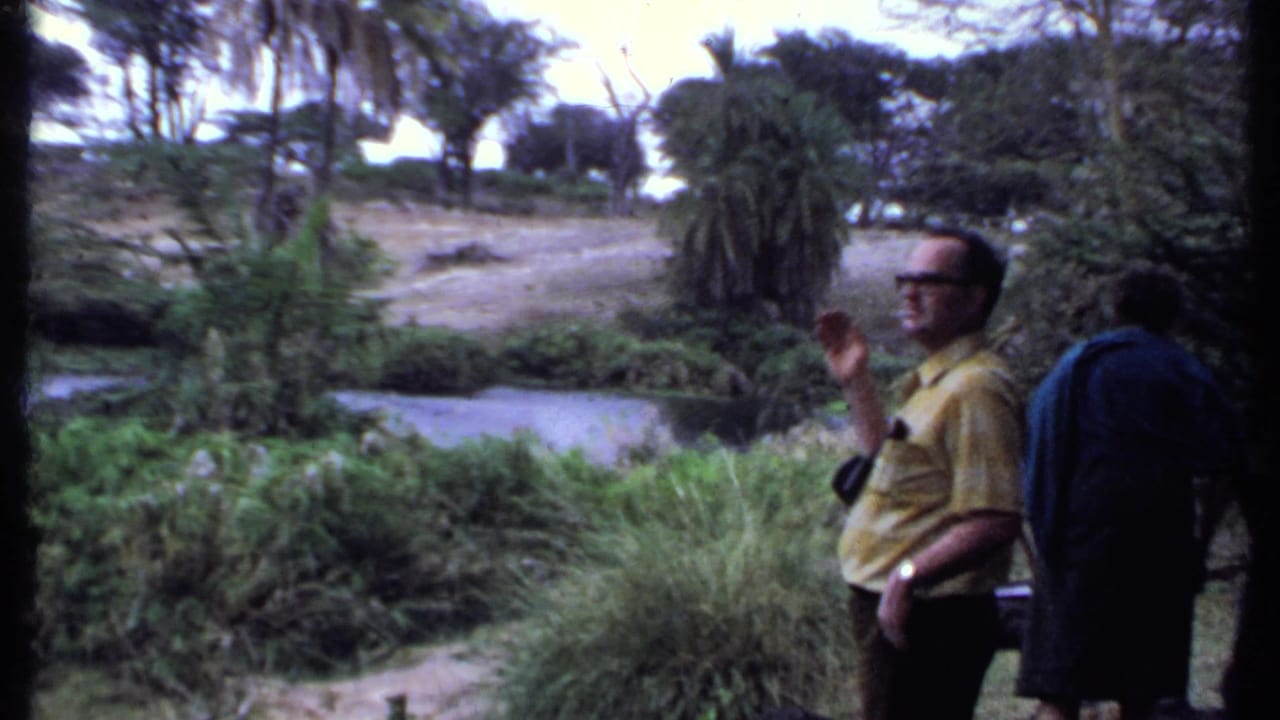 1969 Kenya home-movie frame showing a man in a yellow shirt gesturing by a river, with another visitor partly visible.