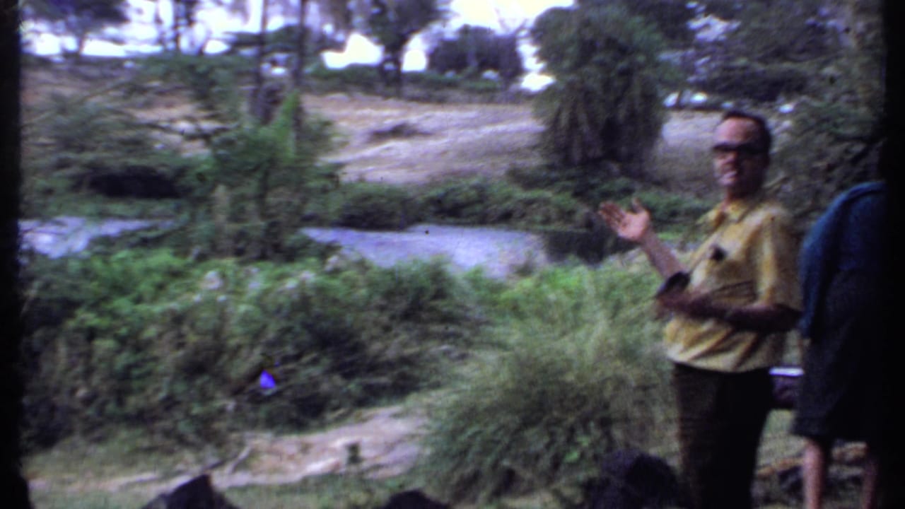 1969 Kenya travel frame with a man in a yellow shirt gesturing beside riverbank plants, trees, and flowing water.