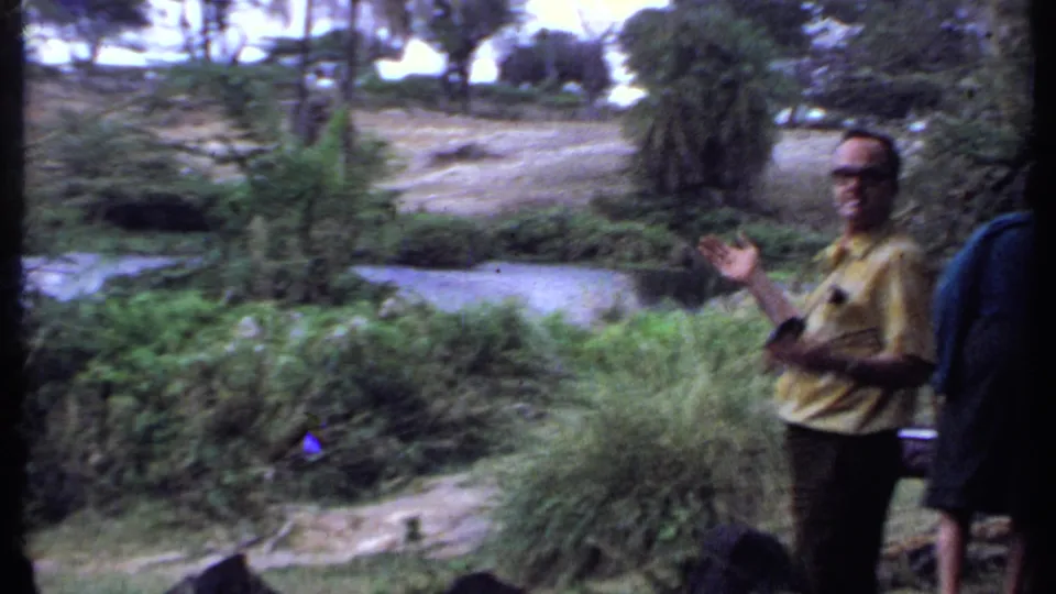 1969 Kenya travel frame with a man in a yellow shirt gesturing beside riverbank plants, trees, and flowing water.