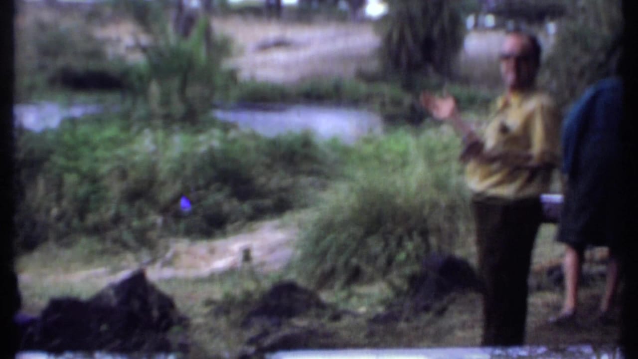Man in a yellow shirt stands near foliage and water with another adult partly visible.