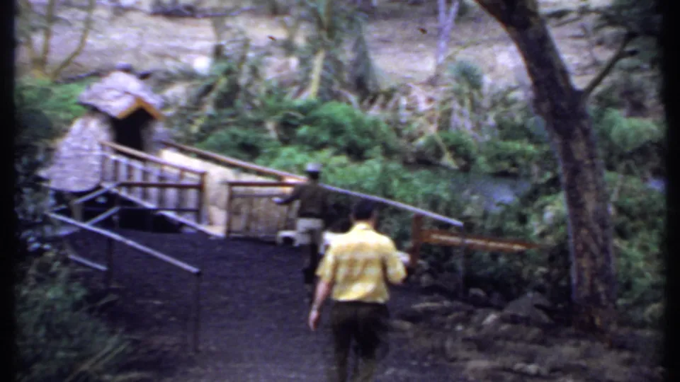 1969 Kenya 8mm frame showing a man in a yellow shirt walking along a path toward a small wooden bridge beside greenery and water.