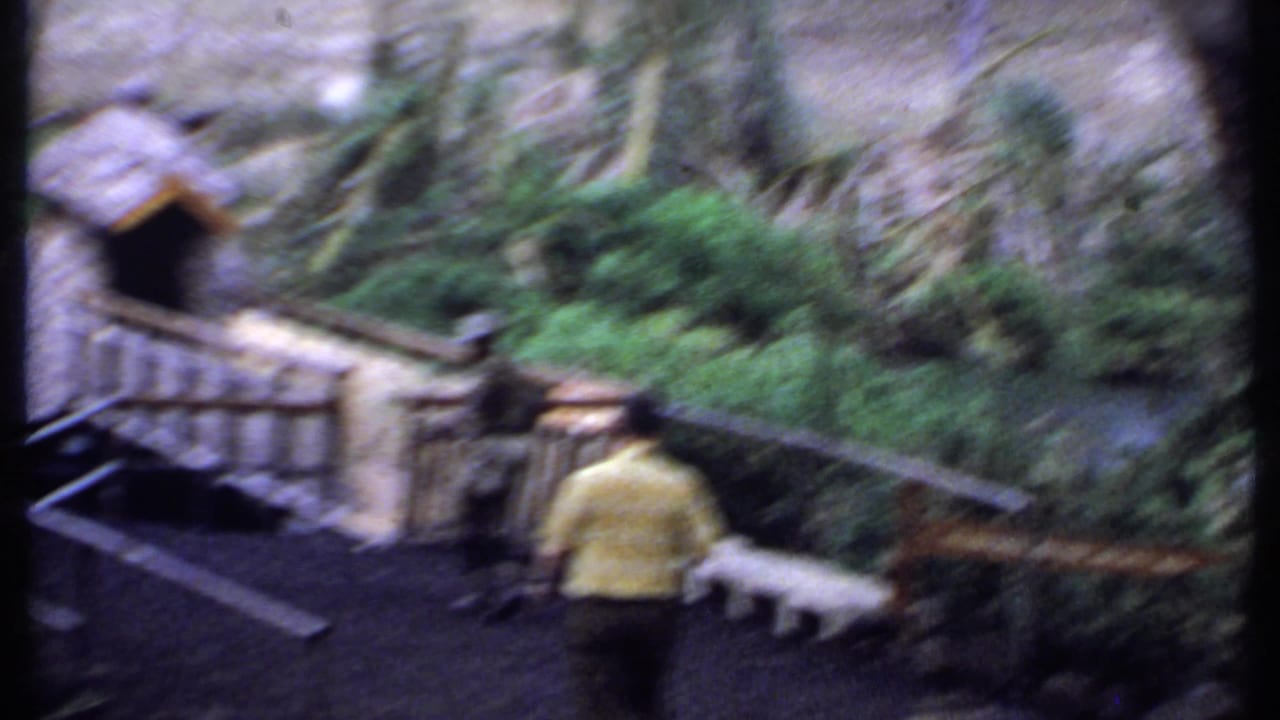 Soft 1969 Kenya home-movie frame with a man in a yellow shirt near a small bridge, stream, hut-like structure, and dense greenery.