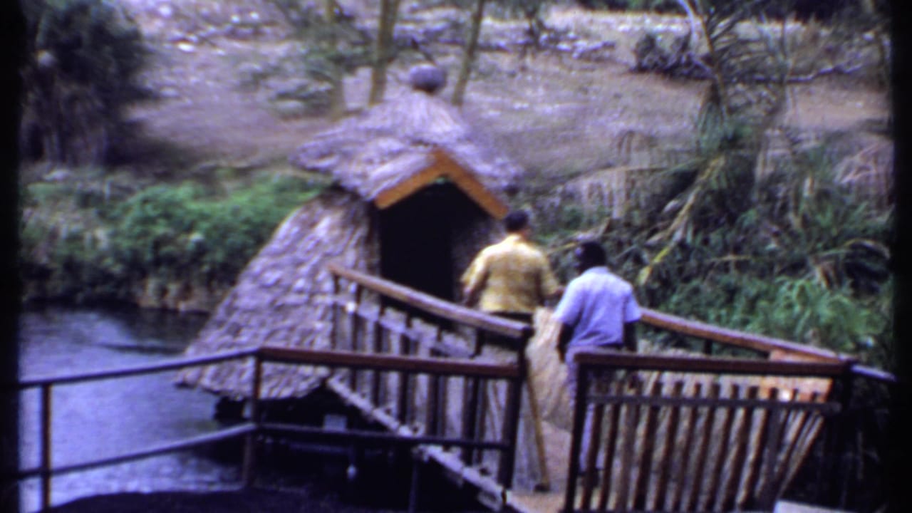 1969 Kenya frame of two men walking across a wooden bridge toward a thatched hut beside running water.
