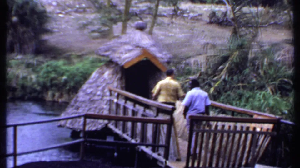1969 Kenya frame of two men walking across a wooden bridge toward a thatched hut beside running water.