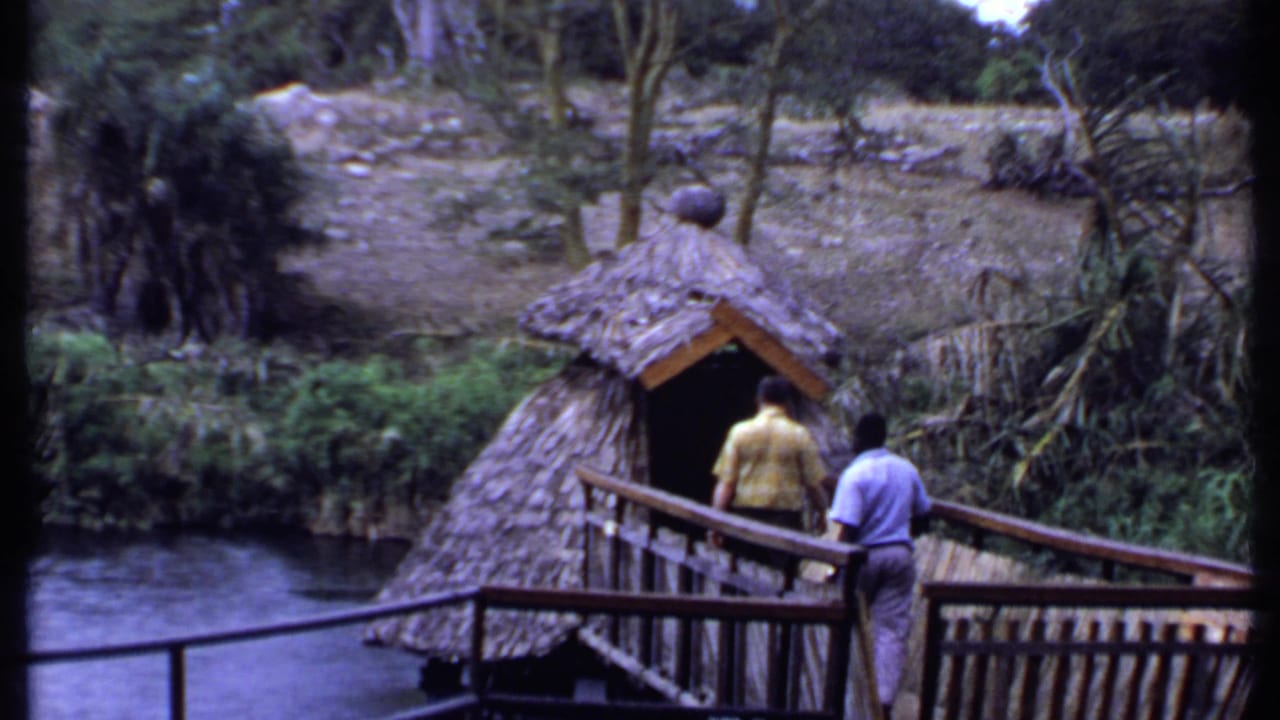 1969 Kenya 8mm frame with two men on a wooden bridge beside a thatched hut, water, and tropical vegetation.