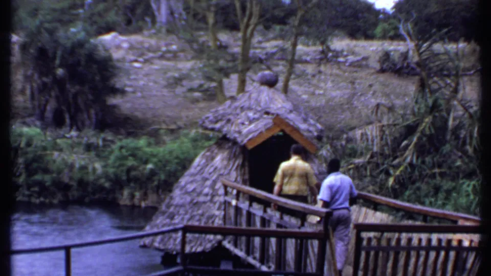 1969 Kenya 8mm frame with two men on a wooden bridge beside a thatched hut, water, and tropical vegetation.