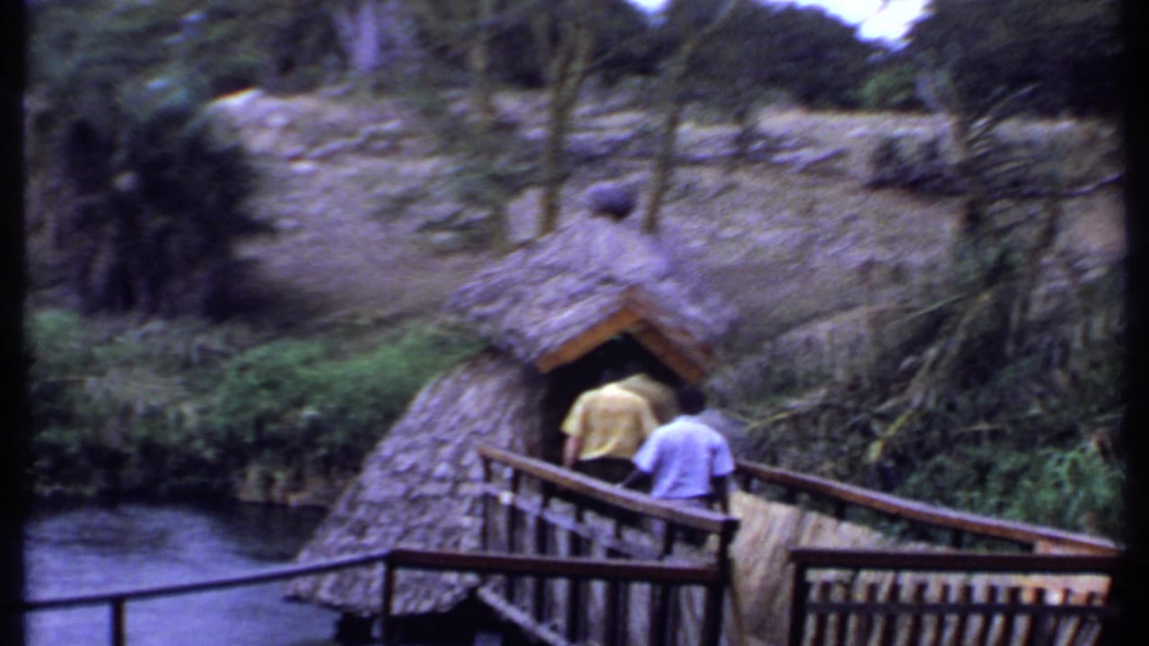 1969 Kenya home-movie frame showing two men approaching a thatched hut from a wooden bridge over water.