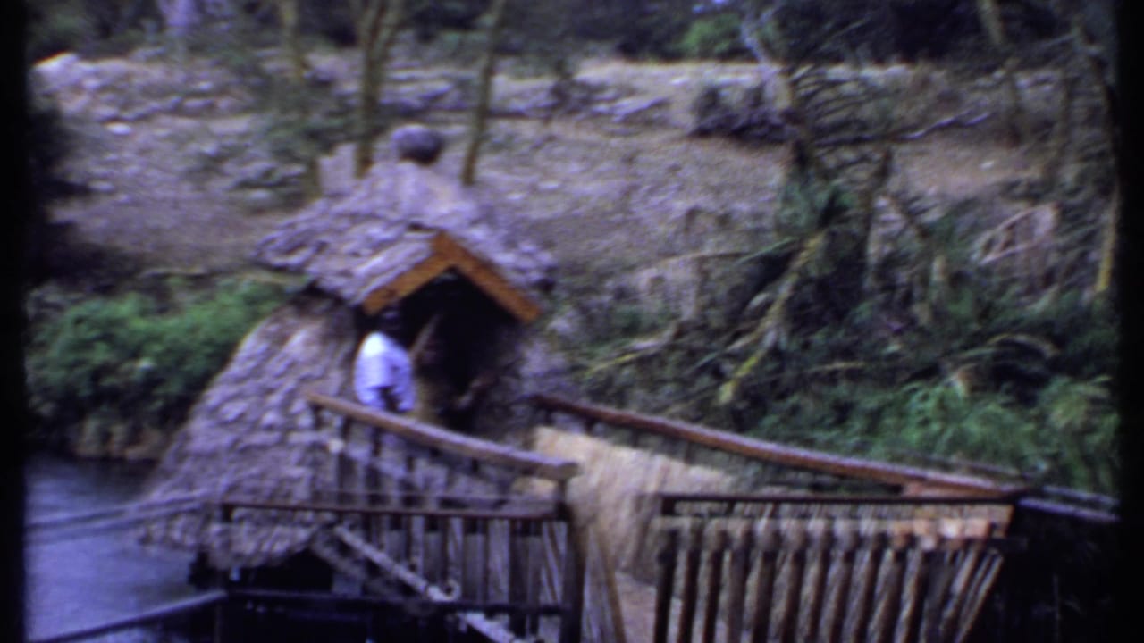 1969 Kenya home-movie frame of a man in a white shirt at a thatched structure near wooden railings, water and planted banks.