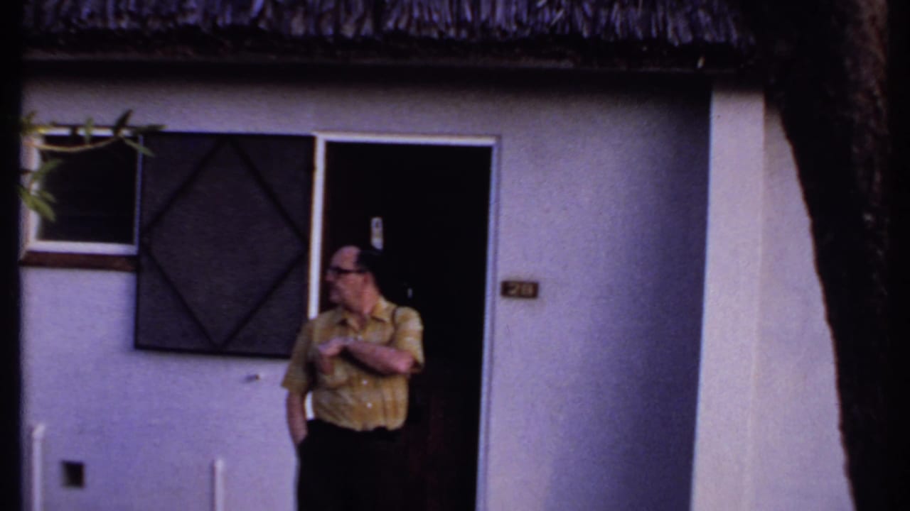 Kenya 1969 home-movie frame of a man in a yellow shirt standing outside a thatched-roof building.