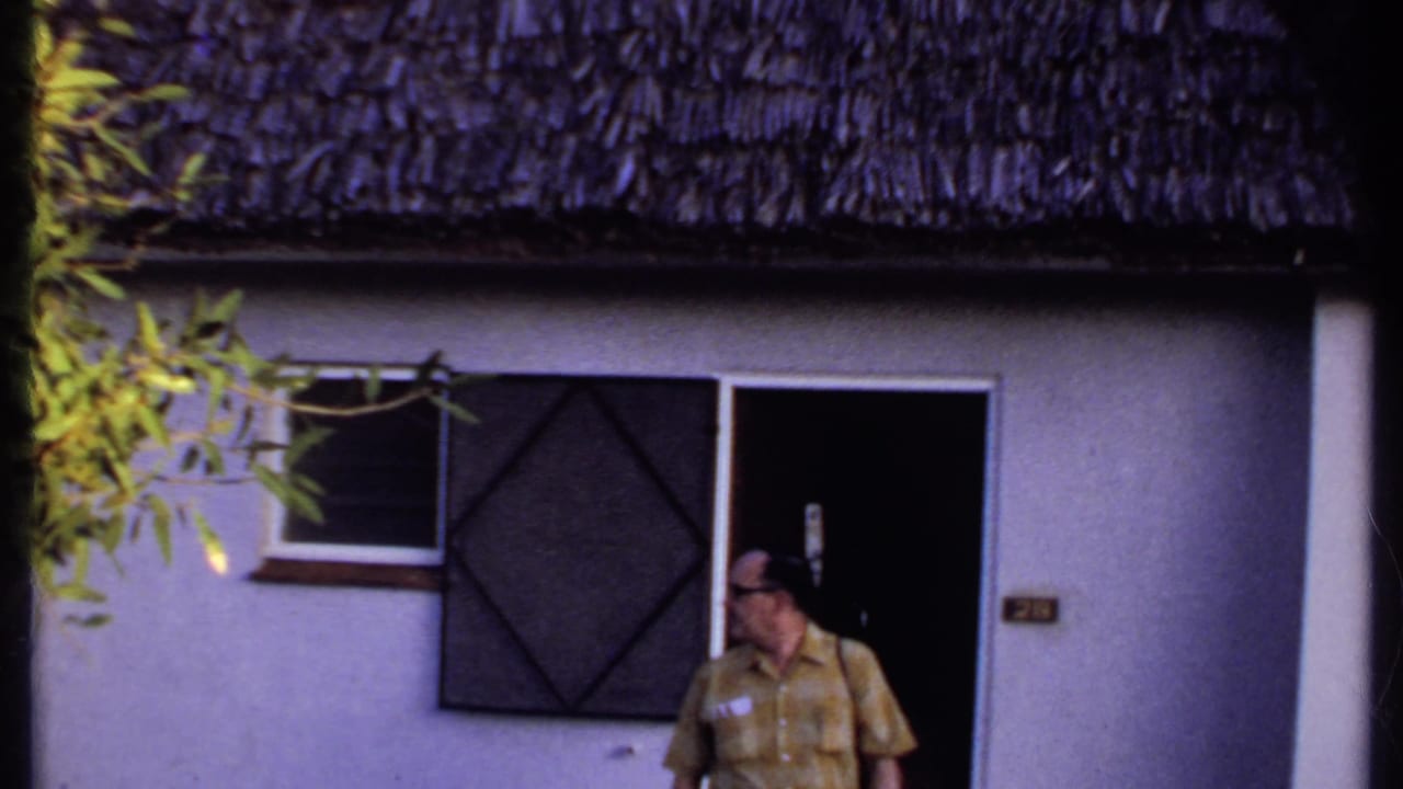Kenya 1969 frame showing a man outside a light-colored thatched-roof building, partly framed by nearby foliage.
