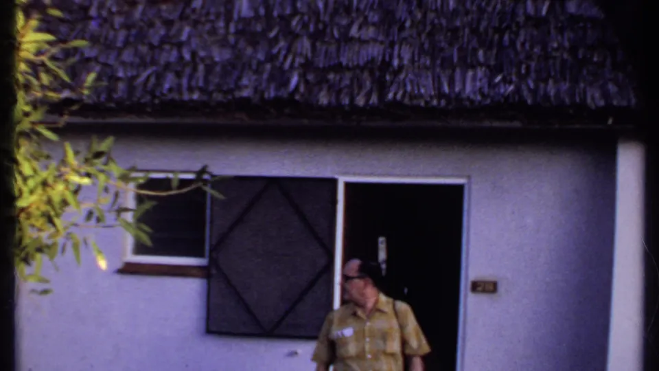 Kenya 1969 frame showing a man outside a light-colored thatched-roof building, partly framed by nearby foliage.