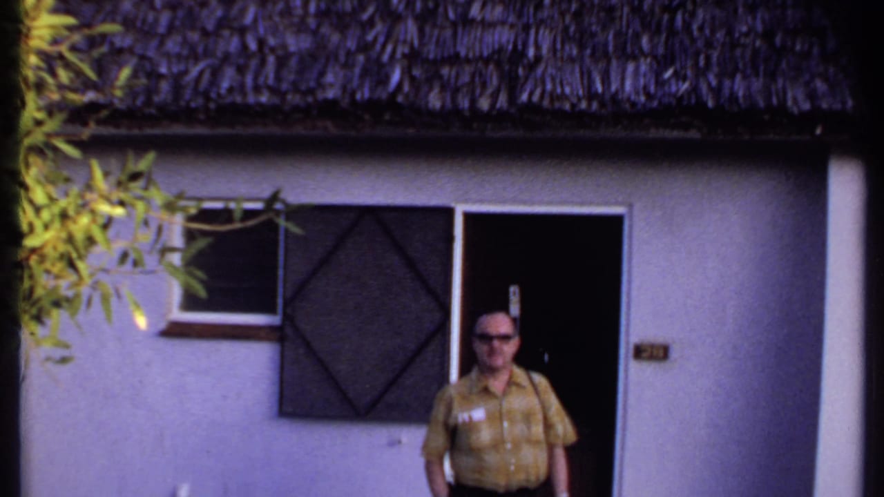 Man in sunglasses standing near a doorway beneath a thatched roof.