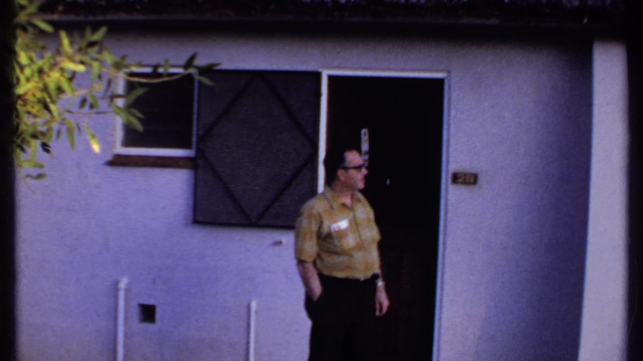 In Kenya, 1969, the man stands partly in the doorway of a white thatched-roof building and looks to the side.
