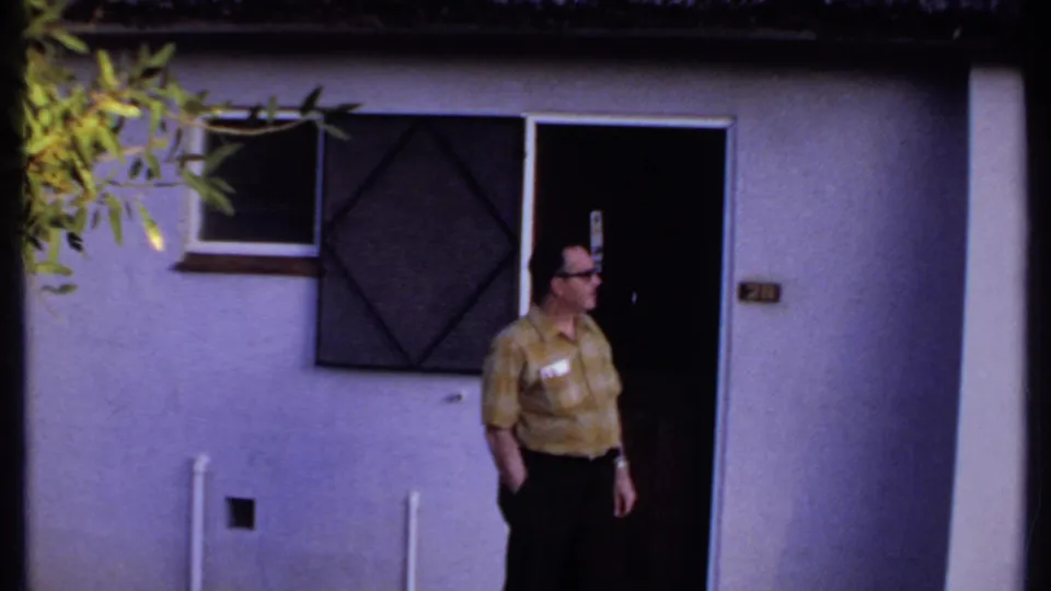 In Kenya, 1969, the man stands partly in the doorway of a white thatched-roof building and looks to the side.