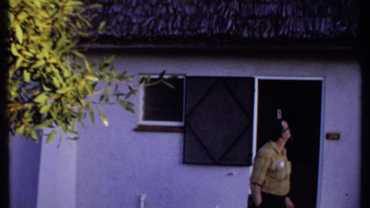 In Kenya, 1969, the man turns near the dark entrance of a white building with a thatched roof and screened window.