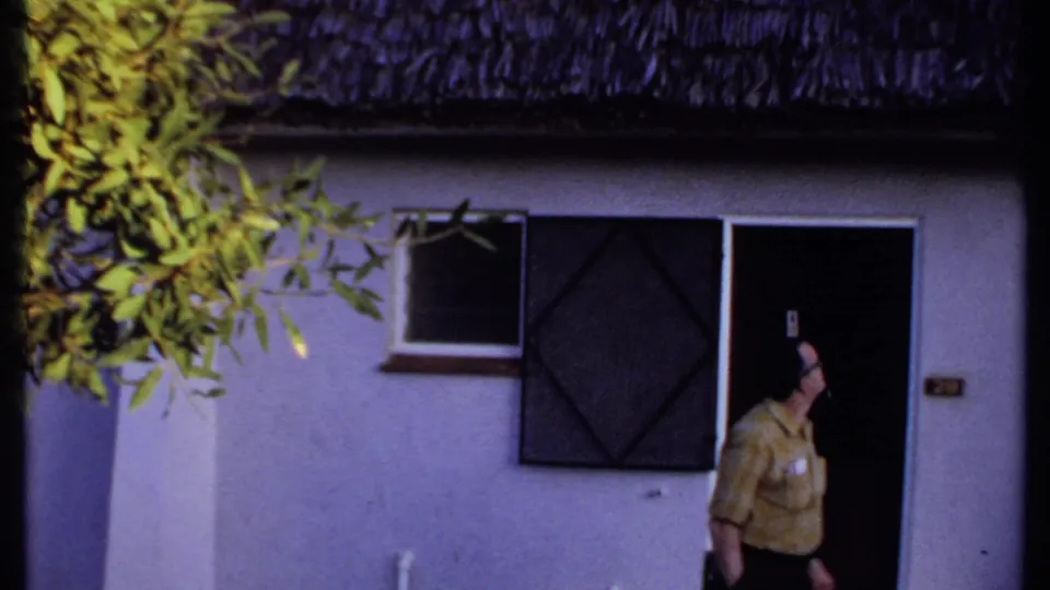 In Kenya, 1969, the man turns near the dark entrance of a white building with a thatched roof and screened window.