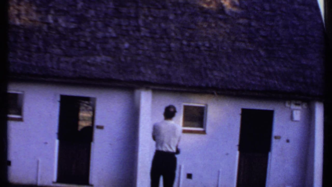 In Kenya, 1969, a rear view shows the man facing a white thatched-roof building with two dark doorways and small windows.